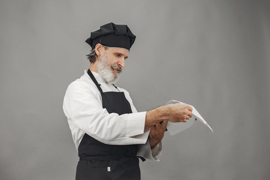 A senior male chef with facial hair wearing a uniform reviews a menu in a studio setting.